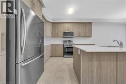 Contemporary kitchen featuring stainless steel appliances, light wood-finish cabinetry, a large kitchen island with an undermount sink, white countertops, and neutral-toned tile flooring - 