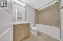 Bathroom featuring a wood-finish vanity with a light-toned countertop, a rectangular mirror, a white toilet, and a white bathtub with light-toned tiled surround - 