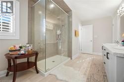 Spacious bathroom featuring a glass-enclosed shower with tiled walls and built-in shelving, dual vanity with undermount sinks and dark hardware, plantation shutters on the window, and light-toned floo - 