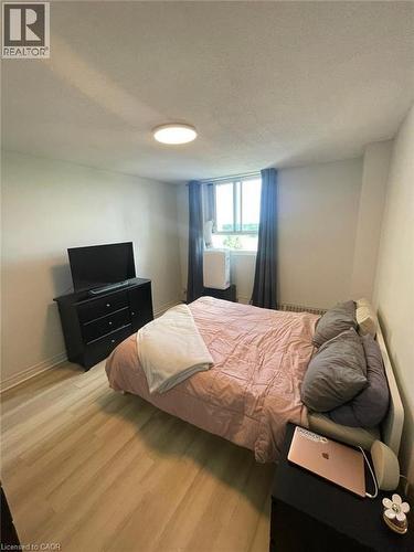 Bedroom featuring wood-finish flooring, a broad window with vertical blinds and dark curtains, and a flush-mount ceiling light fixture - 225 Harvard Place Unit# 1505, Waterloo, ON 