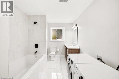 Modern bathroom featuring a white bathtub with matte black fixtures, white large format tile, a wood-finish vanity with a white countertop, and an integrated washer and dryer - 129 St George Street, Kitchener, ON 