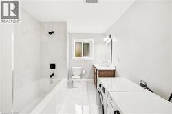 Modern bathroom featuring a white bathtub with matte black fixtures, white large format tile, a wood-finish vanity with a white countertop, and an integrated washer and dryer - 