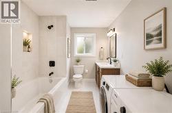 Bathroom featuring a built-in tub with matte black fixtures and a tiled surround, a window providing natural light, a vanity with wood-finish cabinetry and a single-basin sink, and a stacked washer an - 