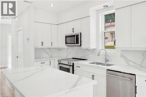 Modern white kitchen featuring an island, white cabinetry, integrated stainless steel appliances, a double basin sink, and wood-finish flooring - 131 St George Street, Kitchener, ON 