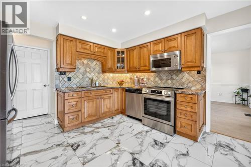 Kitchen featuring light wood cabinetry, stainless steel appliances, patterned backsplash, granite countertops, and white marble-look tile flooring - 15 Cheval Drive, Grimsby, ON - Indoor Photo Showing Kitchen