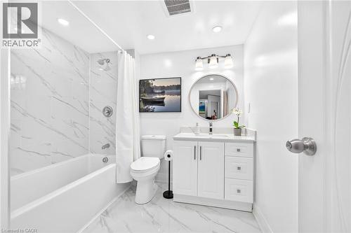 Bathroom featuring white marble-look wall and floor tile, a white vanity with a light-colored countertop, and a circular mirror with an overhead three-light fixture - 15 Cheval Drive, Grimsby, ON - Indoor Photo Showing Bathroom