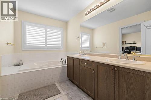 Bathroom featuring a built-in tub with tile surround, white plantation shutters, a dual sink vanity with wood-finish cabinetry, and a large mirror - 4670 Thomas Alton Boulevard, Burlington, ON 