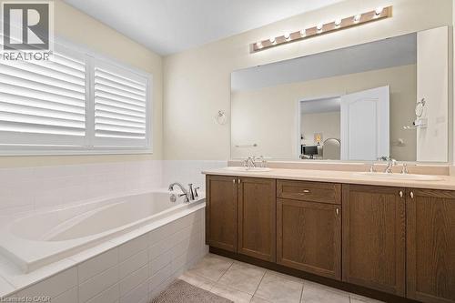 Built-in soaking tub with white tile surround, accompanied by a double vanity featuring wood-finish cabinetry, integrated sinks, a light-toned countertop, and an expansive wall-mounted mirror - 4670 Thomas Alton Boulevard, Burlington, ON 
