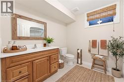 Bathroom featuring a light wood-finish vanity with a white countertop and integrated sink, a framed mirror, a toilet, a window with a woven shade, and light tile flooring - 