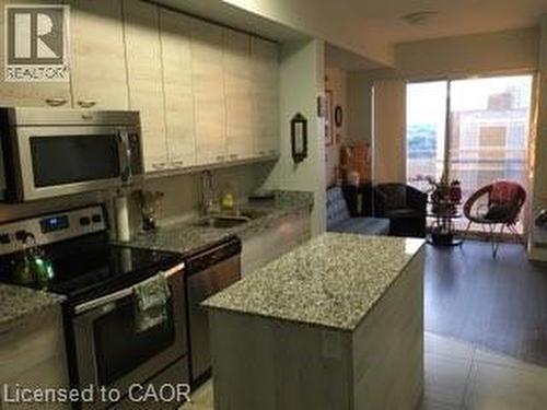 Kitchen island with granite countertop, surrounded by stainless steel appliances, light wood-finish cabinetry, and dark wood-finish flooring extending into a living area with sliding glass doors - 280 Lester Street Unit# 607, Waterloo, ON 