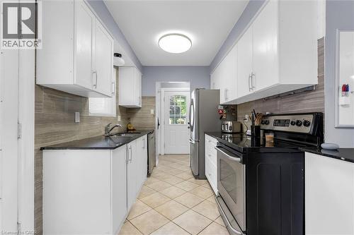 Galley kitchen featuring abundant white cabinetry, dark countertops, a tile backsplash, stainless steel appliances, and tile flooring - 338 East 24Th Street, Hamilton, ON 