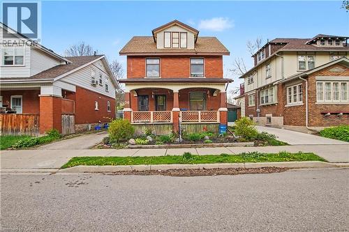 Red brick facade residence featuring a covered front porch with decorative railing, dormer window, and a planted front garden bed - 30 Hohner Avenue, Kitchener, ON - Outdoor