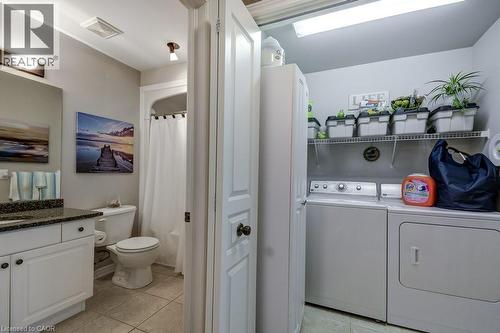 Combined bathroom and laundry area featuring a white vanity with a dark speckled countertop, a toilet, a shower with a white curtain, and a laundry station with a washer, dryer, and wire shelving - 605 Welland Avenue Unit# 21, St. Catharines, ON - Indoor Photo Showing Laundry Room