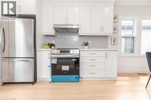 Contemporary kitchen featuring stainless steel appliances, white cabinetry with gold-tone hardware, a subway tile backsplash, light-tone wood-finish flooring, and natural light from multiple windows - 297 Britannia Avenue, Hamilton, ON 