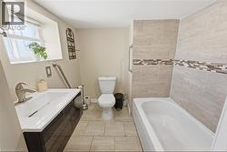 Bathroom featuring a modern vanity with integrated sink, tiled flooring, a standard toilet, and a bathtub with tiled surround and decorative accent band - 