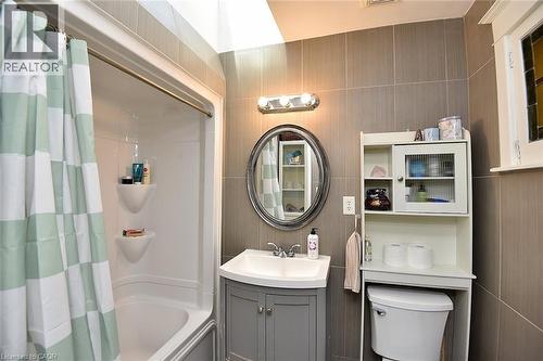 Bathroom featuring a built-in shower/tub combination, gray vanity with integrated sink, oval mirror, wall-mounted light fixture, and floor-to-ceiling tile - 56 Barons Avenue N, Hamilton, ON 