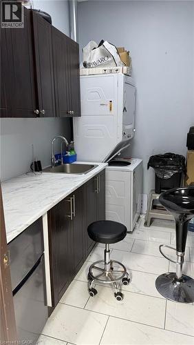 Compact utility area featuring a stacked washer and dryer, dark wood-finish cabinetry, a stainless steel sink with a gooseneck faucet, light-toned countertop, and light-toned tile flooring - 9 Beaumaris Drive Unit# B4, Brampton, ON 