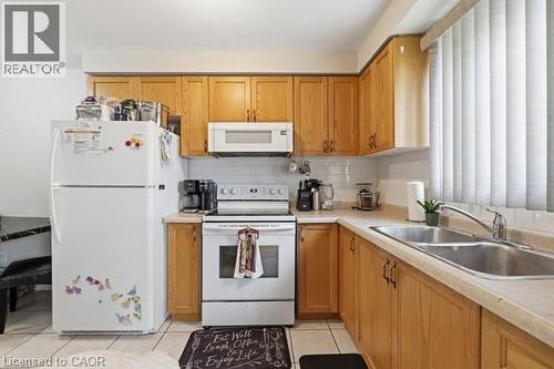 Kitchen featuring wood-finish cabinetry, white appliances, a double basin stainless steel sink, tile backsplash, and light-toned countertops - 16 Harrison Avenue, Hamilton, ON 