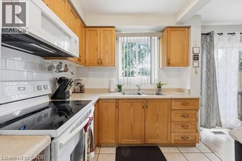 Kitchen featuring wood-finish cabinetry, a white subway tile backsplash, a double basin sink with window, white appliances, and light-toned floor tiling - 16 Harrison Avenue, Hamilton, ON 