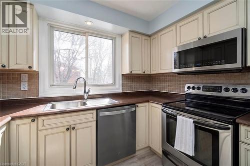Corner kitchen featuring cream-colored cabinetry, stainless steel appliances, a tile backsplash, and a large window above the sink - 121 Morgan Avenue Unit# 7, Kitchener, ON 