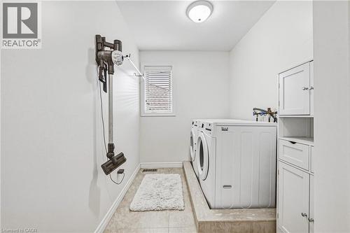 Dedicated laundry area featuring front-loading appliances on a raised platform, built-in white cabinetry with various storage options, a window with blinds, and recessed ceiling lighting - 20 Degrow Crescent, Binbrook, ON 