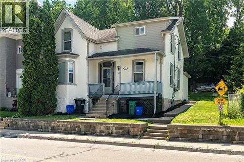 Traditional architecture featuring a covered front porch with a blue railing, bay window, and a stone retaining wall - 66 Queen Street W, Cambridge, ON 