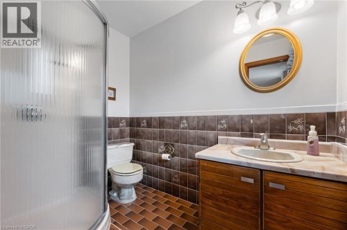 Bathroom featuring a shower stall with fluted glass doors, a vanity with wood-finish cabinetry, a marble-patterned countertop, a decorative oval mirror, and ceramic floor tiling - 7 Thomson Road, Simcoe, ON 