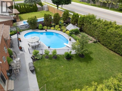 Expansive backyard featuring a kidney-shaped swimming pool with a diving board, surrounding concrete patio, black metal fencing, and mature perimeter landscaping - 7 Thomson Road, Simcoe, ON 