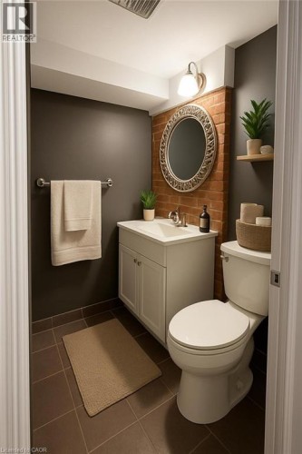 Powder room featuring a white single-sink vanity, tile flooring, a decorative mirror, exposed brick accent wall, and a single wall-mounted light fixture - 947 Main Street E, Hamilton, ON 