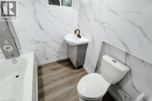 Bathroom featuring white marbled wall panels, wood-finish flooring, white soaking tub with a multi-function shower panel, and a white toilet - 78 Hillyard Street, Hamilton, ON 