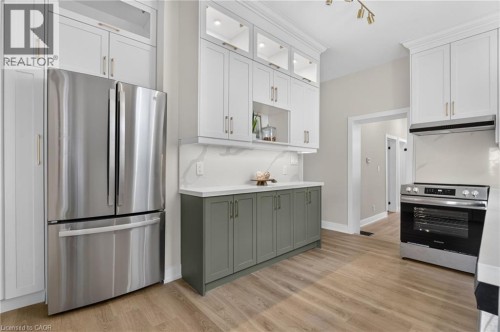 Contemporary kitchen featuring stainless steel appliances, light wood-finish flooring, white upper cabinetry with glass-front accents, and contrasting sage green lower cabinetry - 118 Cathcart Street, Hamilton, ON 