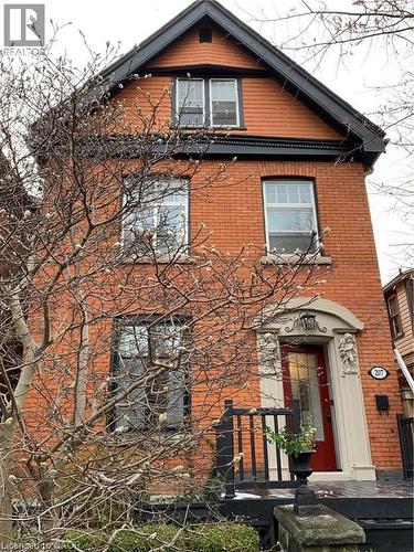 Charming brick Colonial featuring a gabled roofline with dark architectural detailing, multi-pane double-hung windows, and a covered entry portico with red door and black iron railings - 207 Charlton Avenue W, Hamilton, ON 