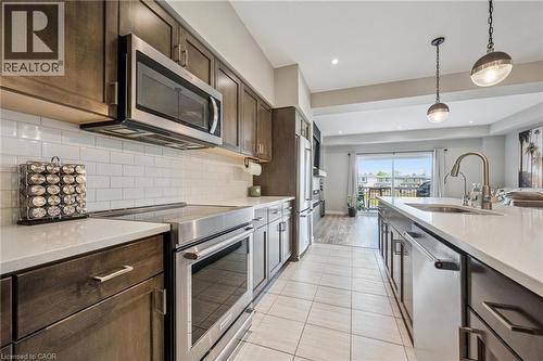 Modern kitchen featuring stainless steel appliances, dark wood-finish cabinetry, a white subway tile backsplash, light stone-finish countertops, and an island with an undermount sink and pendant light - 907 Robert Ferrie Drive, Kitchener, ON 