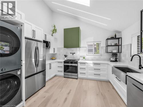Modern kitchen featuring white cabinetry, stainless steel appliances, a paneled green range hood, wood-finish flooring, and a stainless steel farmhouse sink - 13 Swanhurst Boulevard, Mississauga, ON 