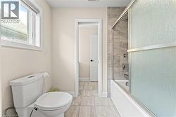 Full bathroom featuring a white ceramic toilet, a window with a white frame, light-toned wall paint, a shower-over-tub combination with a sliding glass door, and a light gray tiled floor - 