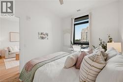Bright bedroom featuring light wood-finish flooring, white walls, and a large window with city architecture visible - 