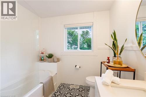 Bathroom featuring a built-in tub with a white surround, single window with white trim, white paneled wainscoting, and a dark and white patterned floor mat - 176 Riverbank Drive, Cambridge, ON 