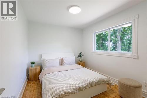 Bedroom featuring light-toned walls, wood-finish flooring, a contemporary ceiling light fixture, a wide window with white trim, and a window framing mature trees - 176 Riverbank Drive, Cambridge, ON 