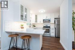 Modern kitchen featuring white cabinetry, subway tile backsplash, stainless steel appliances, wood-finish flooring, and a breakfast bar with white countertop - 