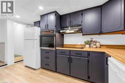 Kitchen featuring dark-toned cabinetry with decorative trim, wood-finish countertops, built-in wall oven, stainless steel range hood, and light wood-finish flooring - 