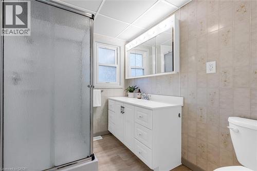 Bathroom featuring a textured glass shower enclosure with chrome framing, a white vanity with integrated sink, a mirrored medicine cabinet with integrated lighting, a window with frosted glass, and ne - 11 Ford Street, Hamilton, ON 