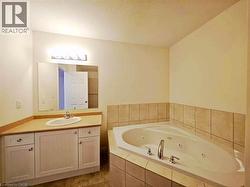 Bathroom featuring a corner jetted tub with tiled surround, a light wood-finish vanity with an integrated sink, a large wall-mounted mirror, and overhead strip lighting - 