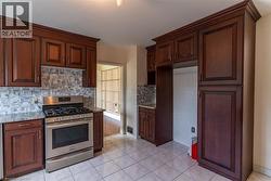 Kitchen featuring abundant dark wood cabinetry, stainless steel range, patterned tile backsplash, light-colored tile flooring, and stone countertops - 