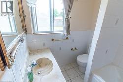 Bathroom featuring a scallop-shell sink integrated into a light-toned countertop, a window with exterior visibility, a toilet, a partial view of a bathtub, and white tiled walls - 