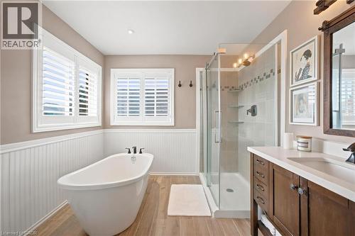 Freestanding soaking tub with dark finish fixtures, wood-finish flooring, glass-enclosed shower with tiled surround, single vanity with white countertop, and white california shutters - 207 Valridge Drive, Ancaster, ON - Indoor Photo Showing Bathroom