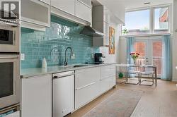Modern kitchen featuring sleek white cabinetry, stainless steel appliances, a vibrant blue subway tile backsplash, light-toned wood-finish flooring, and a large window with transom framing - 