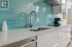 Kitchen featuring a turquoise subway tile backsplash, light-toned countertop, double basin sink with a chrome faucet, stainless steel range hood, and white high-gloss cabinetry - 