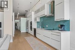 Modern kitchen featuring a teal subway tile backsplash, white flat-panel cabinetry, stainless steel range hood, built-in double oven, and wood-finish flooring - 