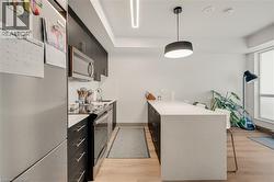 Contemporary kitchen featuring a stone-finish island with seating, stainless steel appliances, dark flat-panel cabinetry, wood-finish flooring, and a linear overhead light fixture - 