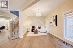 Spacious living area featuring light wood-finish flooring, a contemporary chandelier, large windows with natural light, and a unique geometric wall accent - 